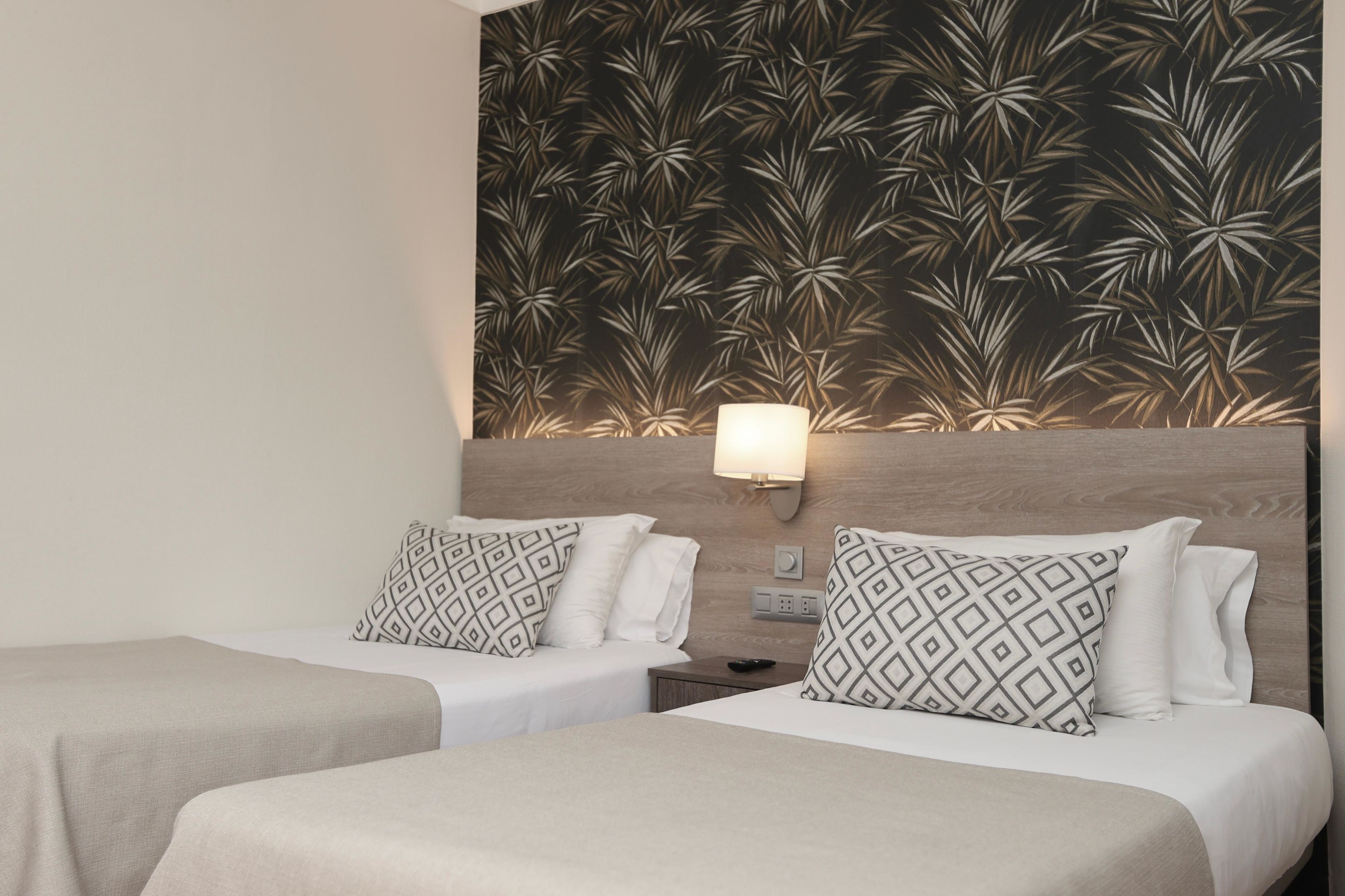 A hotel room with two beds, each with patterned pillows, a nightstand, a wall-mounted lamp, and a decorative wall panel.