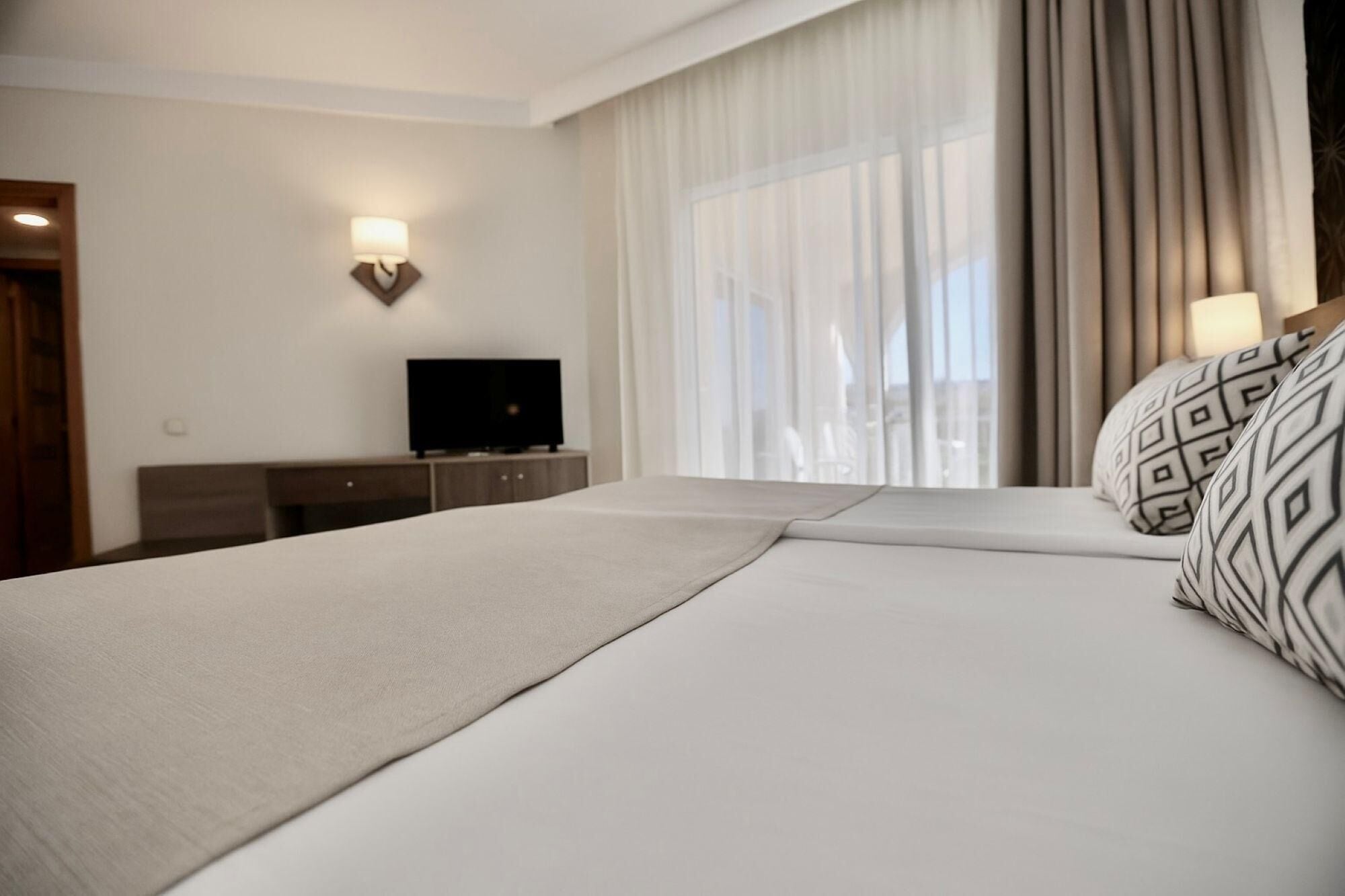 Habitación de hotel con cama, televisor sobre una cómoda de madera, lámpara de pared y ventana con cortinas.