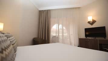 A hotel room with a large bed, a television on a wooden cabinet, a wicker chair, and a window with sheer curtains.