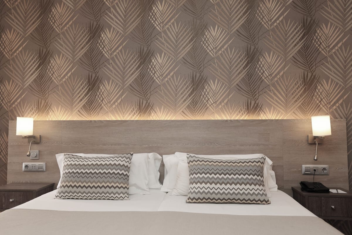 A hotel room with a patterned wallpaper, two bedside lamps, a bed with white and patterned pillows, and a nightstand with a phone and a power outlet.