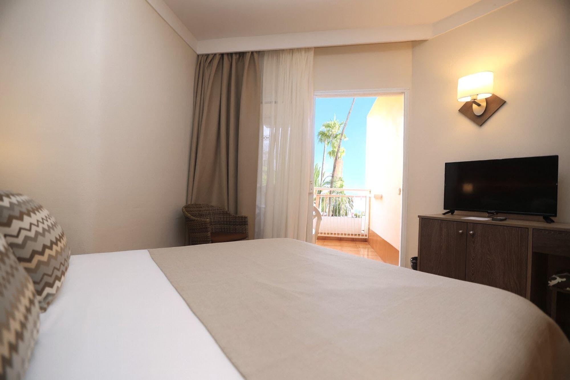 Une chambre d’hôtel avec un grand lit, un téléviseur posé sur une commode en bois, un fauteuil en osier et un balcon offrant une vue sur les palmiers.