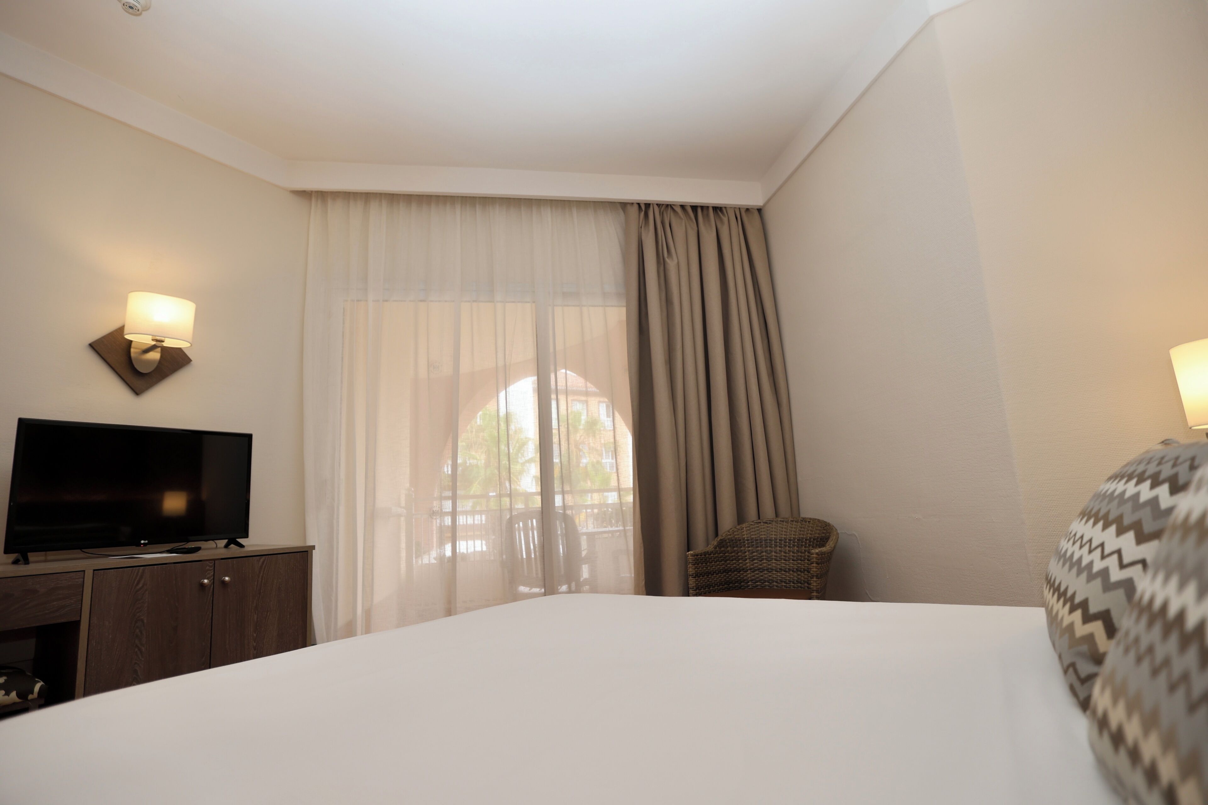 Ein Hotelzimmer mit einem großen Bett, einem Fernseher auf einer Holzkassette, einem Korbstuhl und einem Fenster mit durchscheinenden Vorhängen.