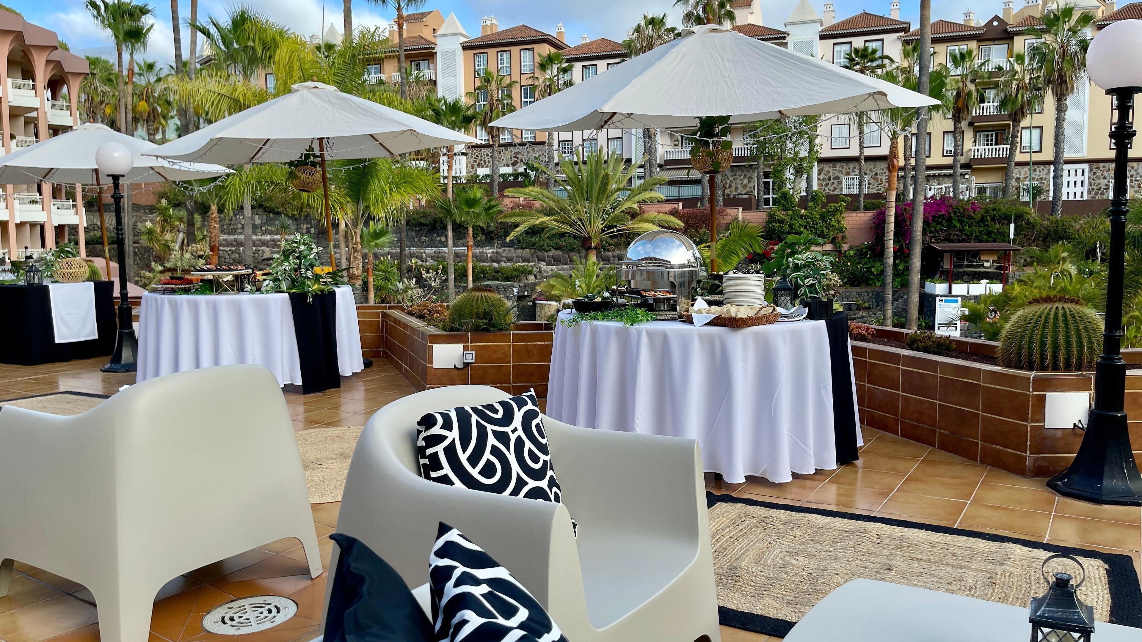 Een terras buiten met witte tafels en stoelen, parasols en uitzicht op een woongebied met palmbomen en een stenen muur.