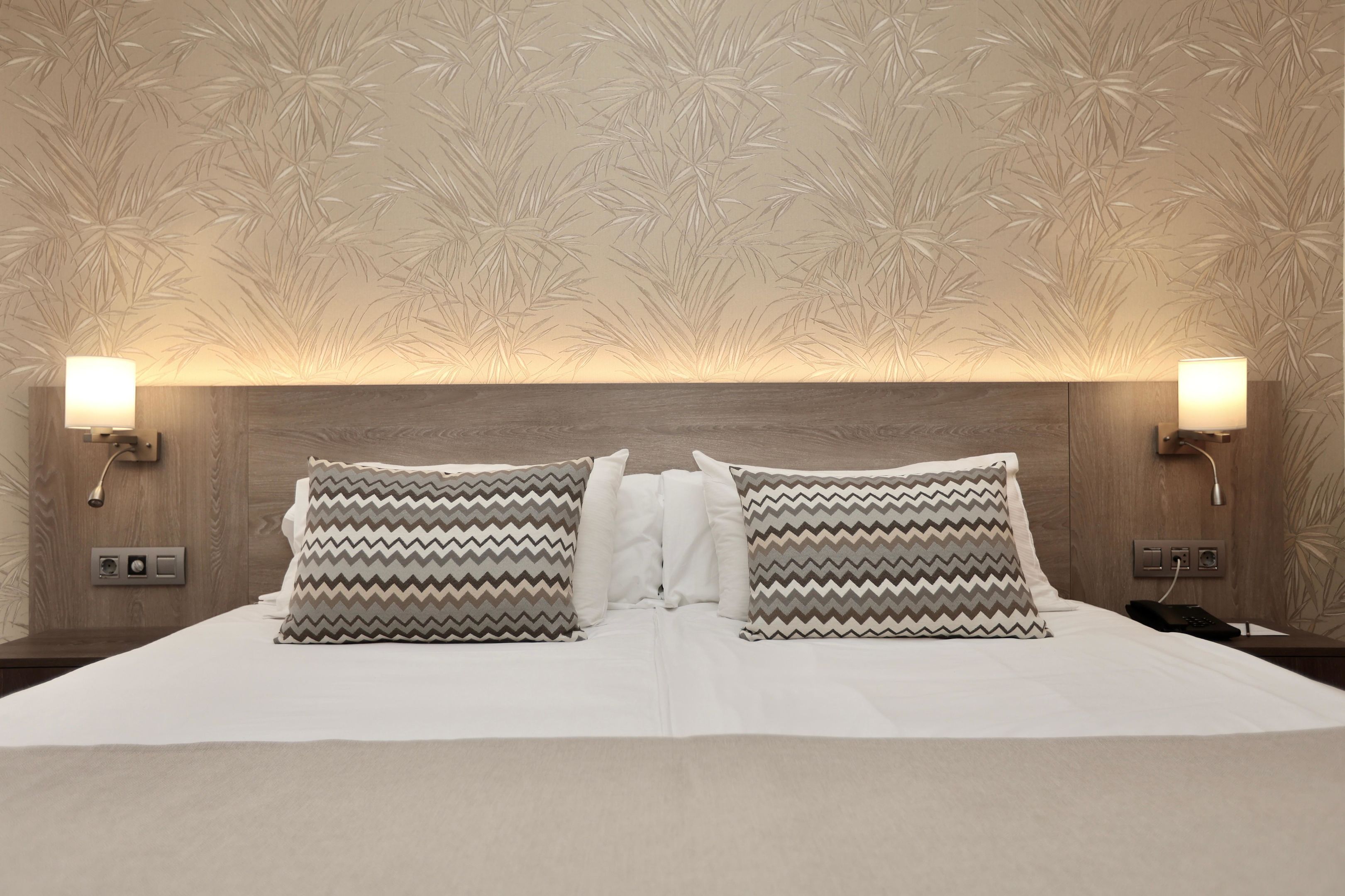 Ein Hotelzimmer mit Holz-Kopfteil, zwei Wandlampen, einem Bett mit weißer Bettwäsche und gemusterten Kissen.