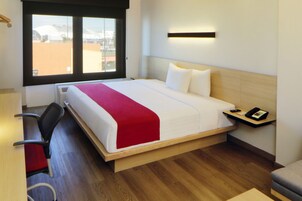 Habitación de hotel con cama, escritorio, silla, mesita de noche y ventana con vista a edificios.