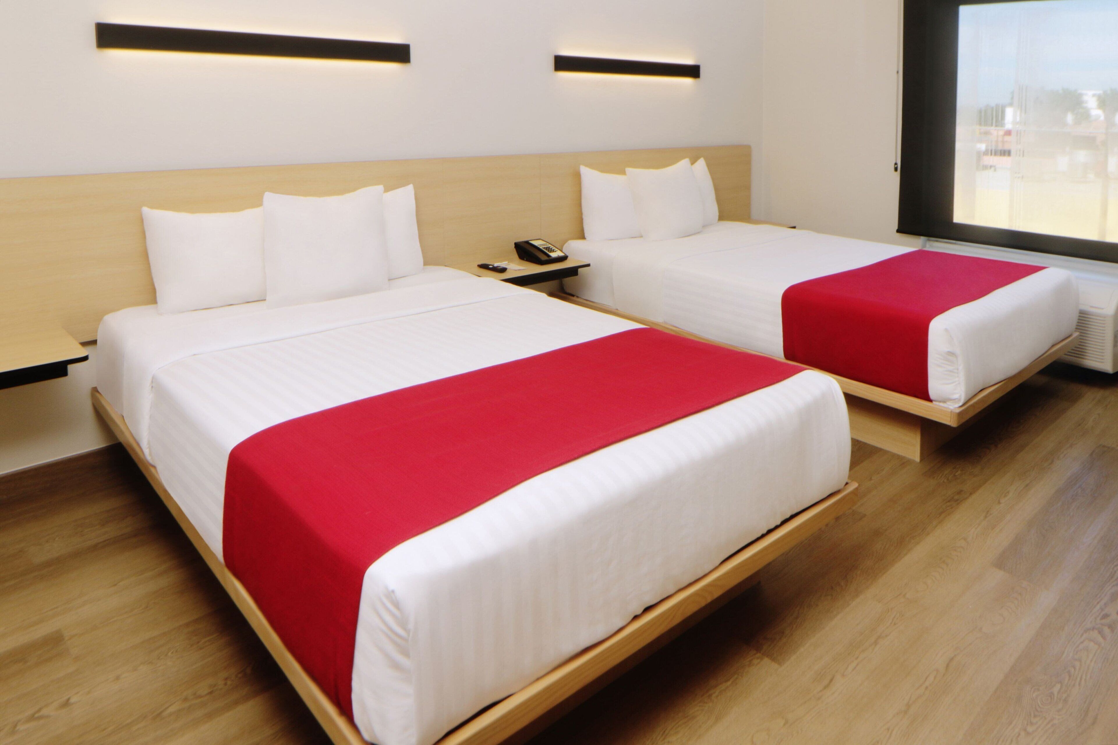 A hotel room with two beds, each with white bedding and a red accent. There are bedside tables with phones and lamps. The room has wooden flooring and a large window with a view.