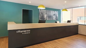 Een receptiebalie met een bordje 'cityexpress hotels', twee zwarte stoelen, een groot abstract kunstwerk aan de muur en een geel hanglamp.