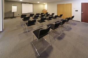 Un salón de conferencias con filas de sillas negras, una pizarra y una pantalla de proyección.
