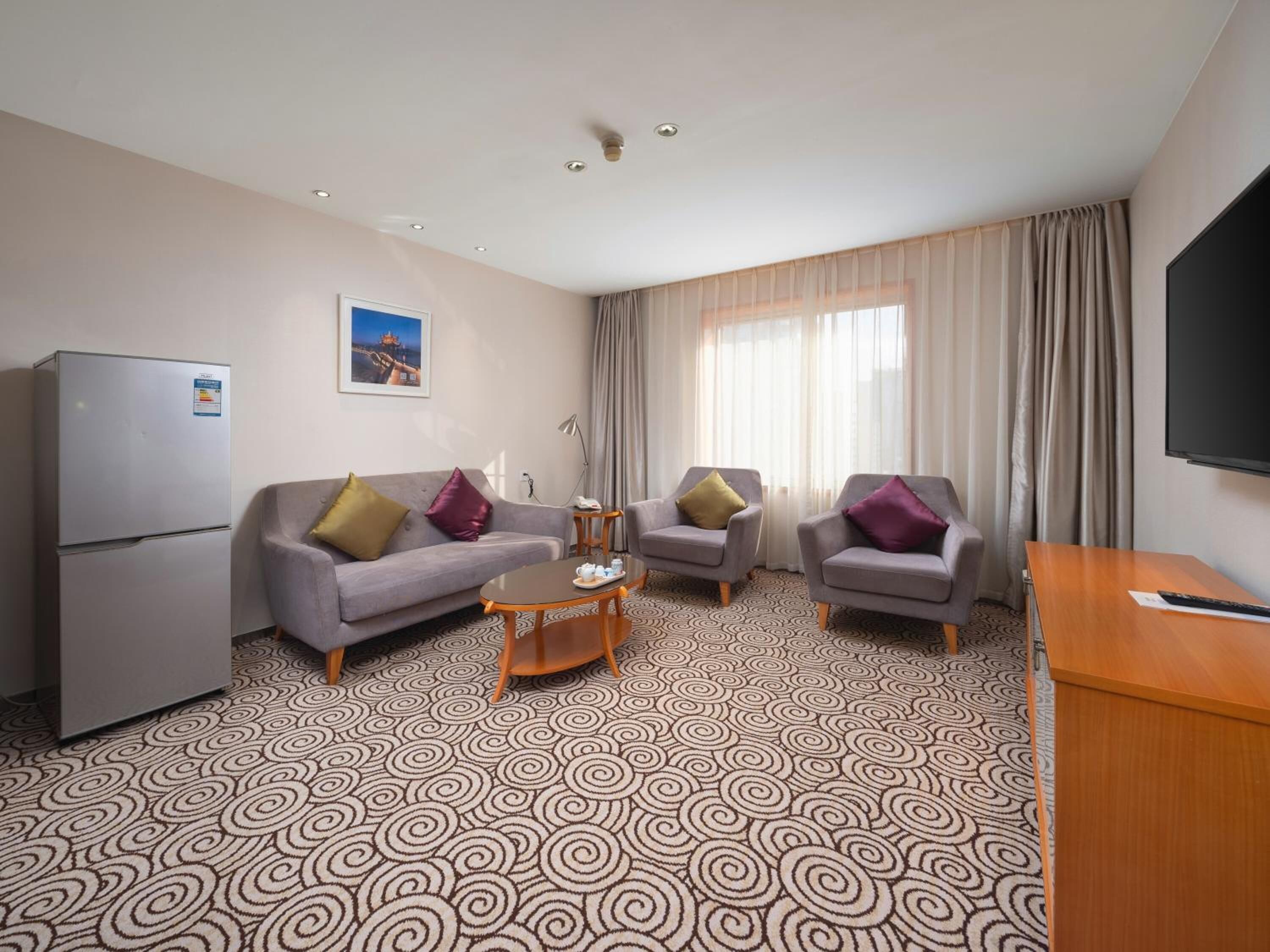 Habitación de hotel con un sofá gris, dos sillones, una mesita de centro, televisión y refrigerador.