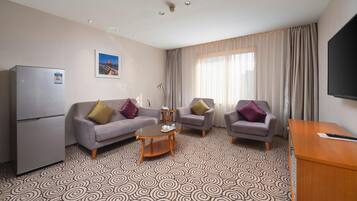 Habitación de hotel con un sofá gris, dos sillones, una mesita de centro, televisión y refrigerador.