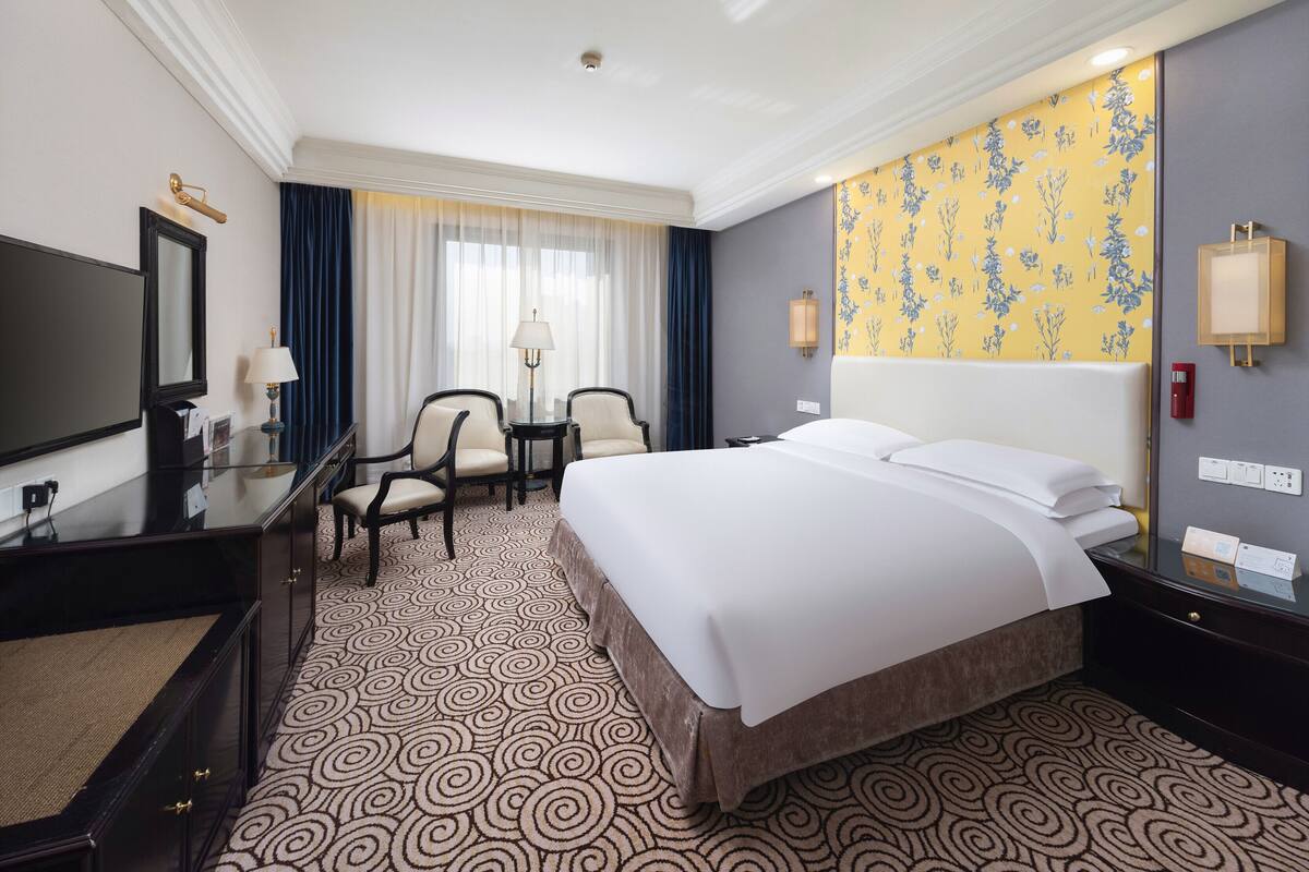 Une chambre d’hôtel avec un grand lit, deux fauteuils, un bureau avec une lampe, une télévision et une moquette à motifs.