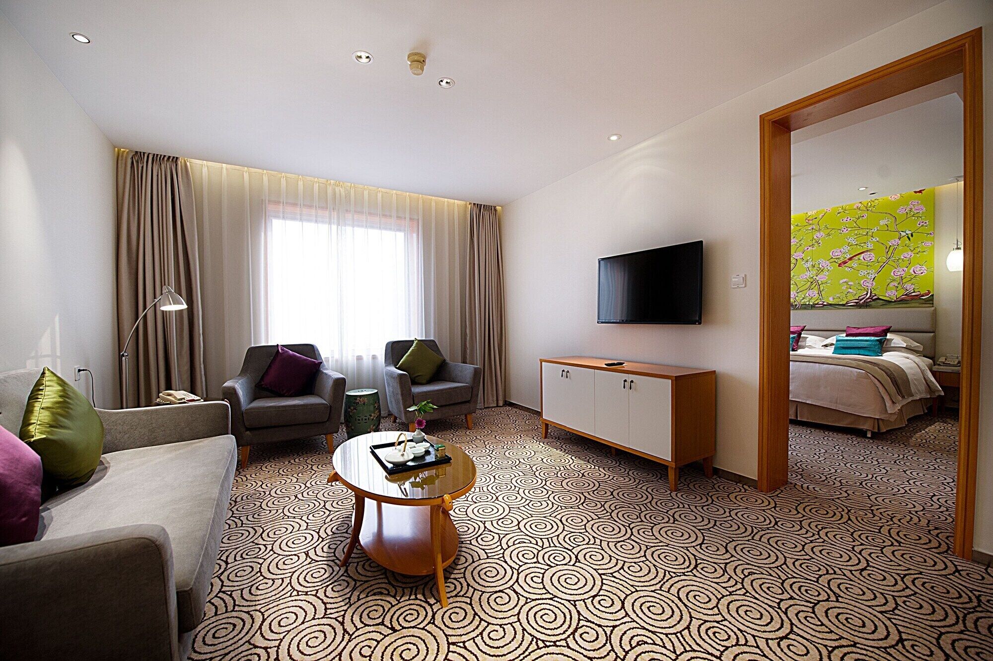 Hotelkamer met een bank, twee fauteuils, een salontafel, een televisie, een kast en een bed met bloemenbehang.