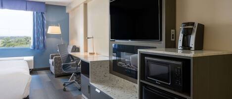 Habitación de hotel con cama, escritorio, silla, televisión, cafetera y microondas, y vista al exterior.
