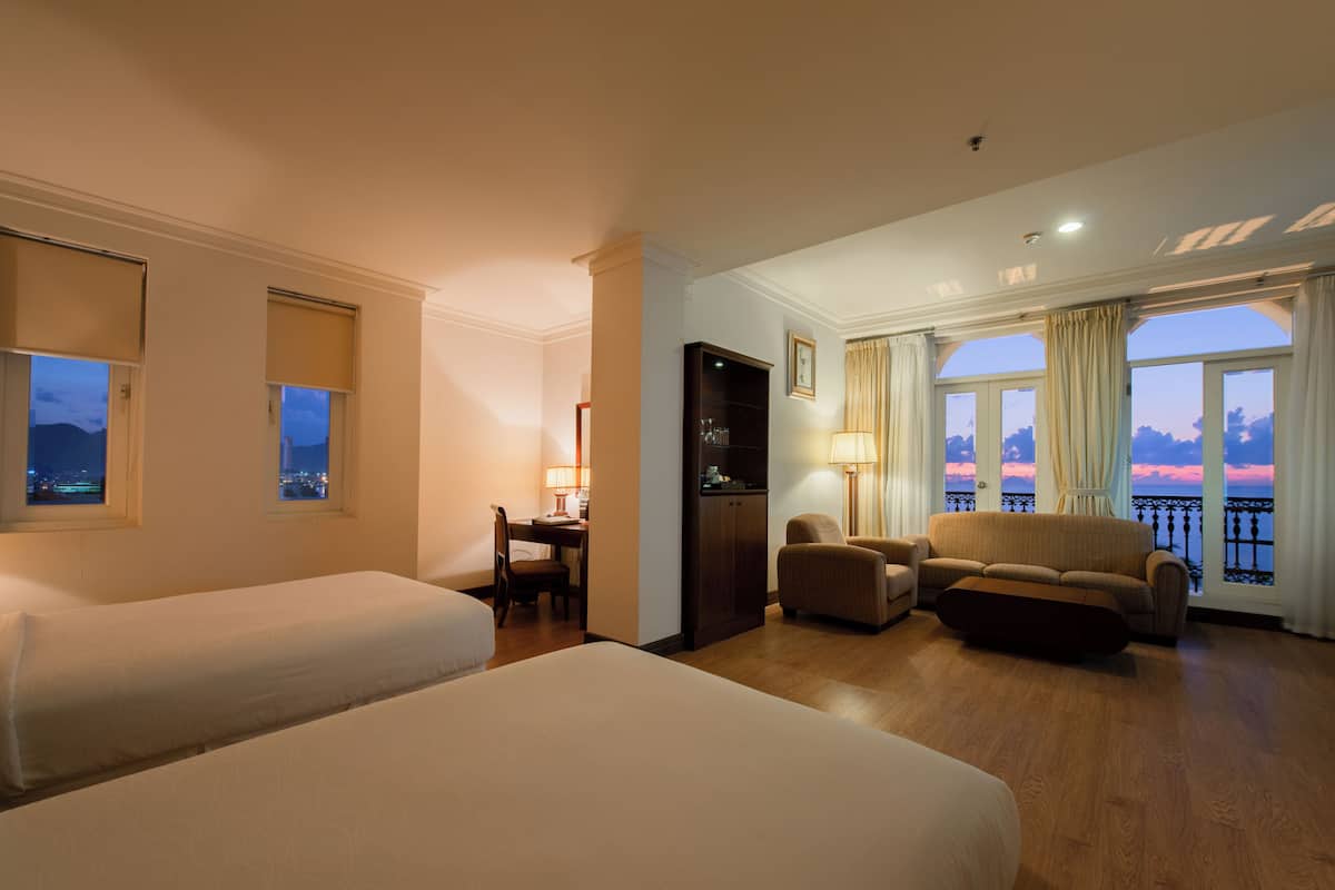 A hotel room with two beds, a desk, a sofa, and a view of the sunset.