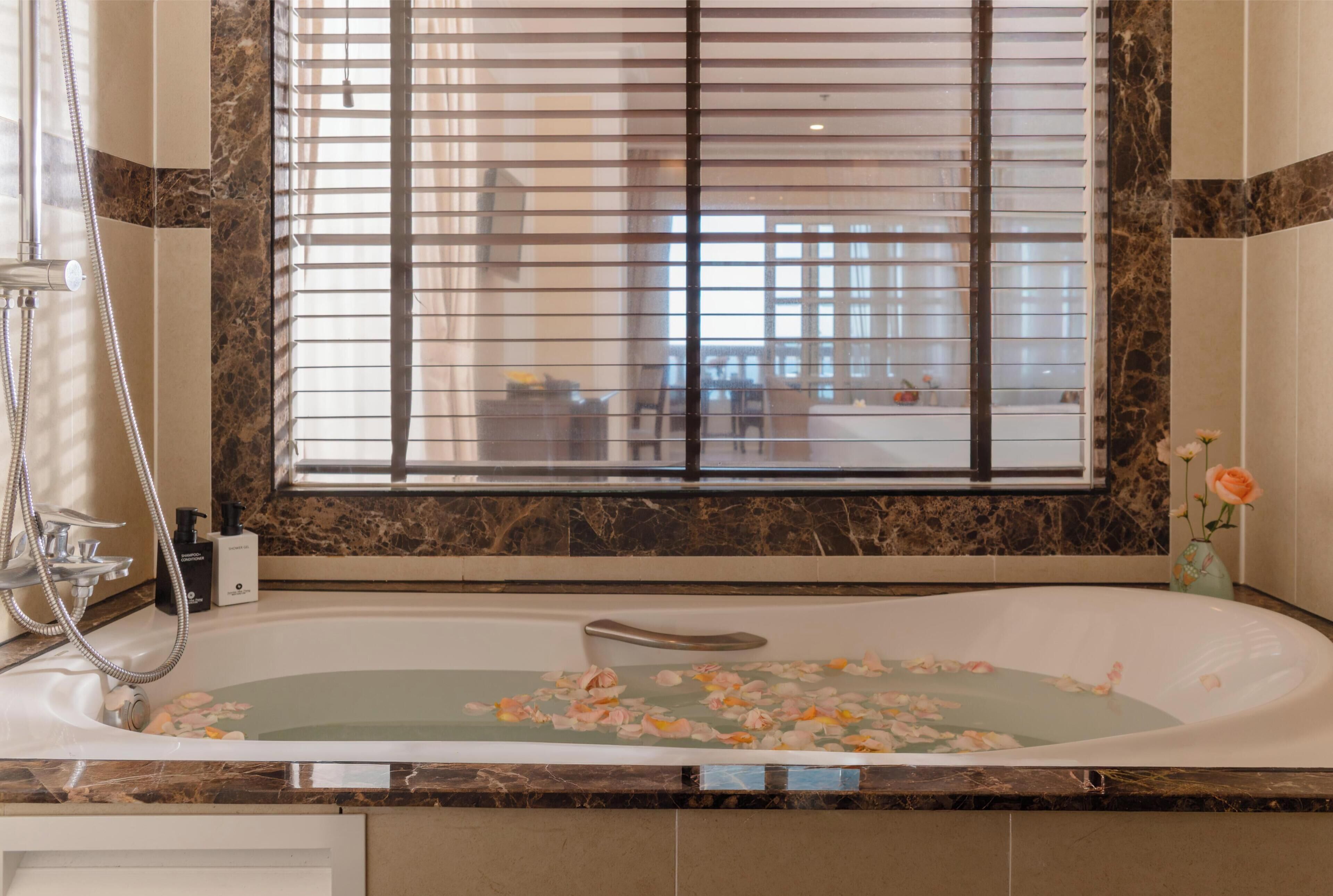 Un baño con una bañera llena de pétalos de rosa, ducha y ventana con persianas.