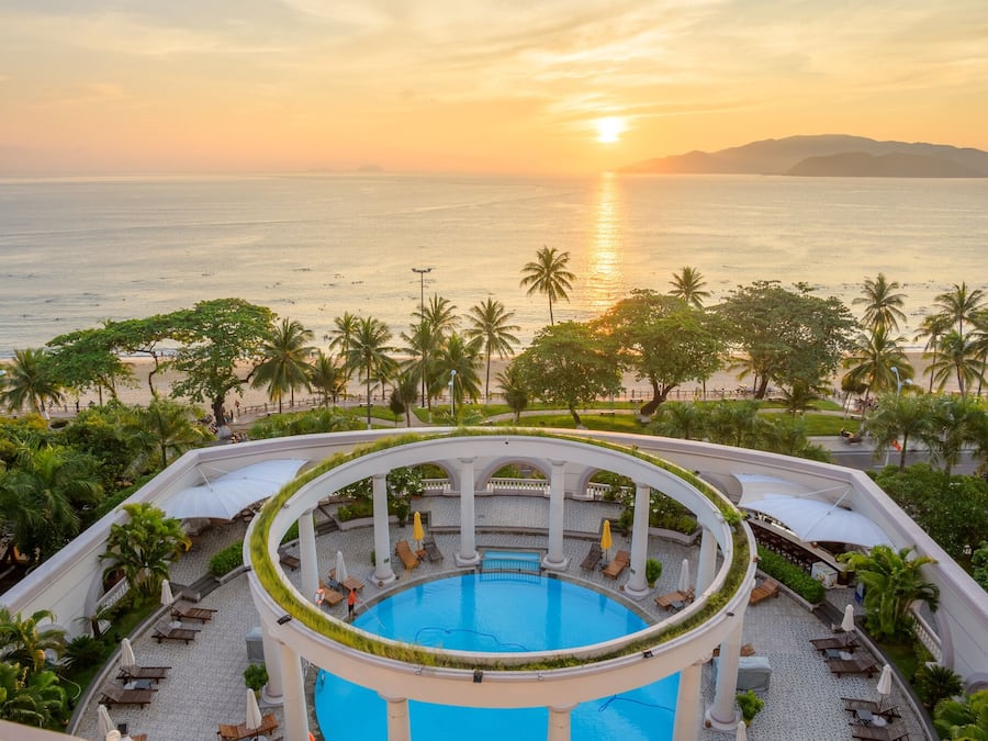 A circular pool with a view of the sunset over the sea, surrounded by palm trees and lounge chairs.