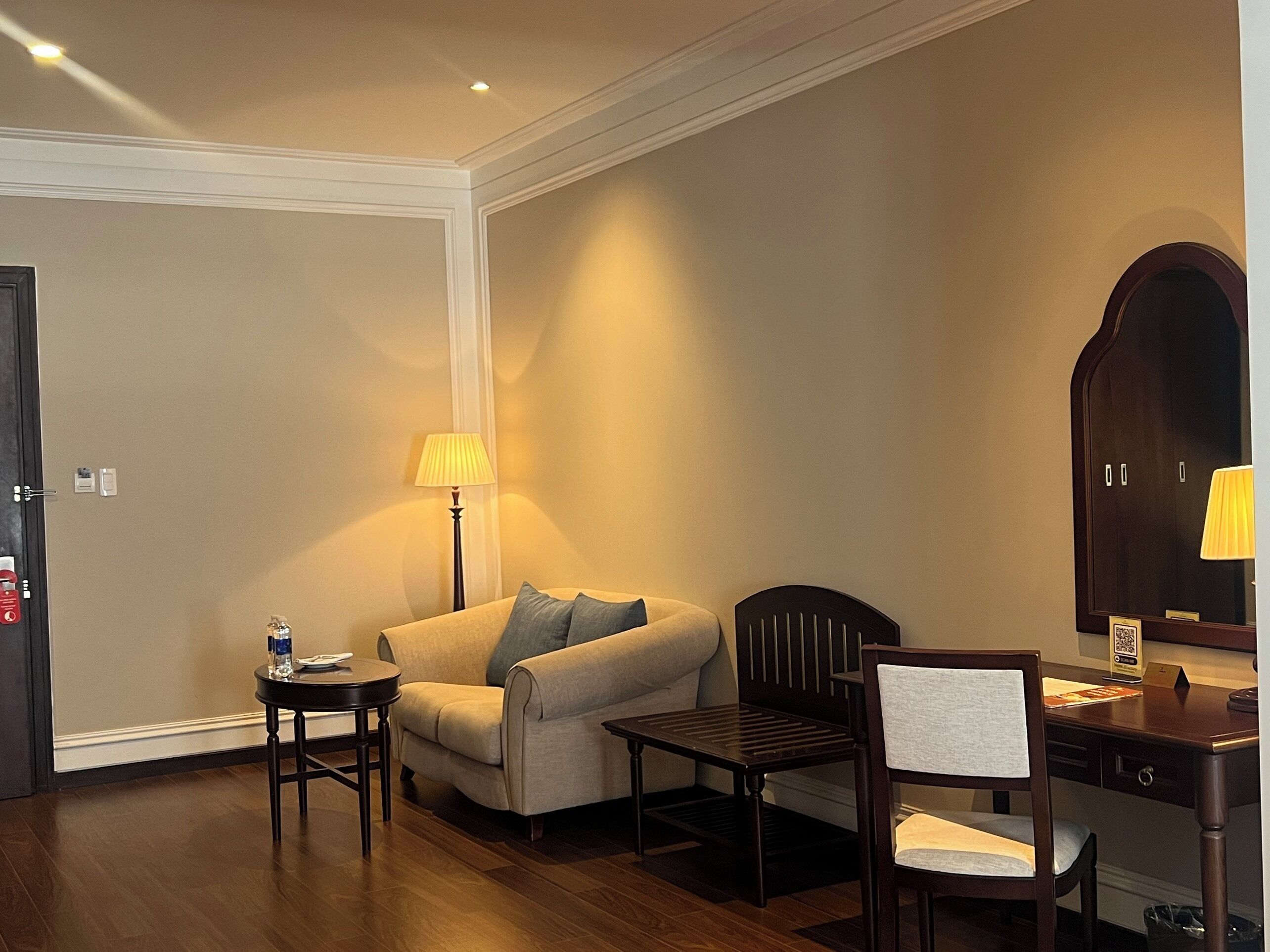 A hotel room with a sofa, a desk, a chair, a lamp, and a wardrobe.