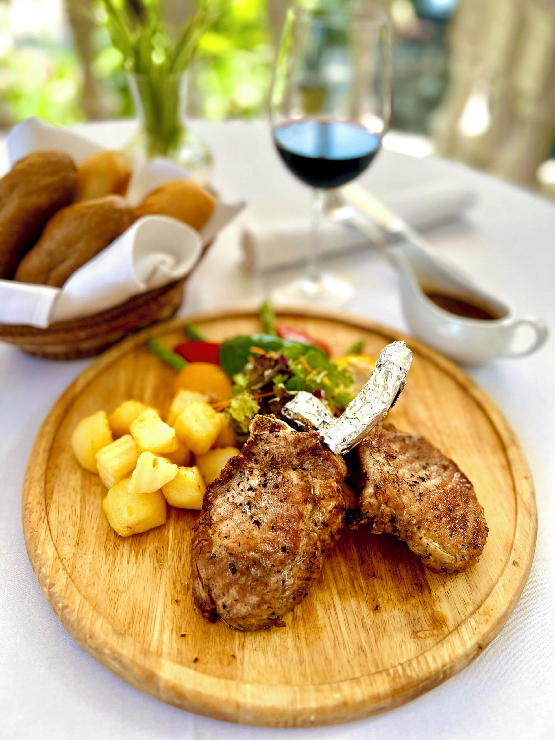 A wooden board with grilled meat, pineapple chunks, and a salad on a table with a glass of red wine and bread basket in the background.
