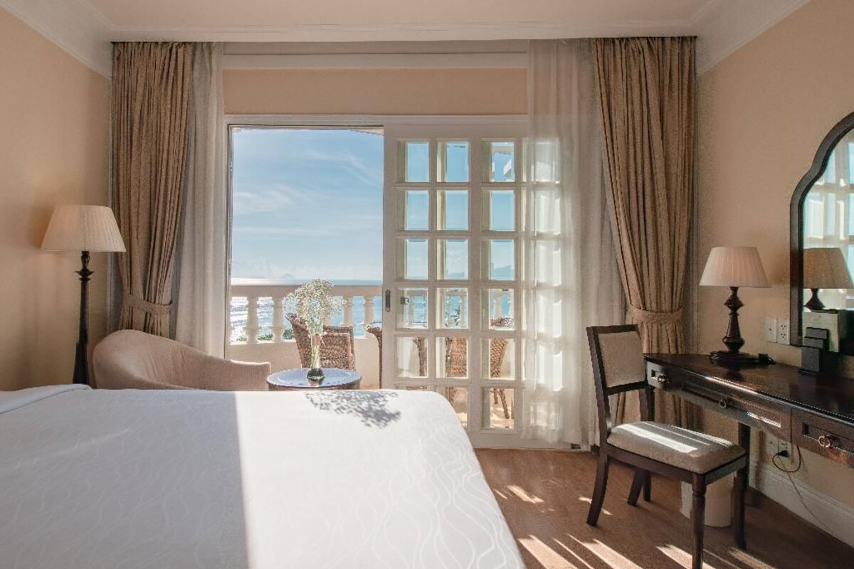 A hotel room with a bed, two chairs, a desk, and a view of the sea through a large window.