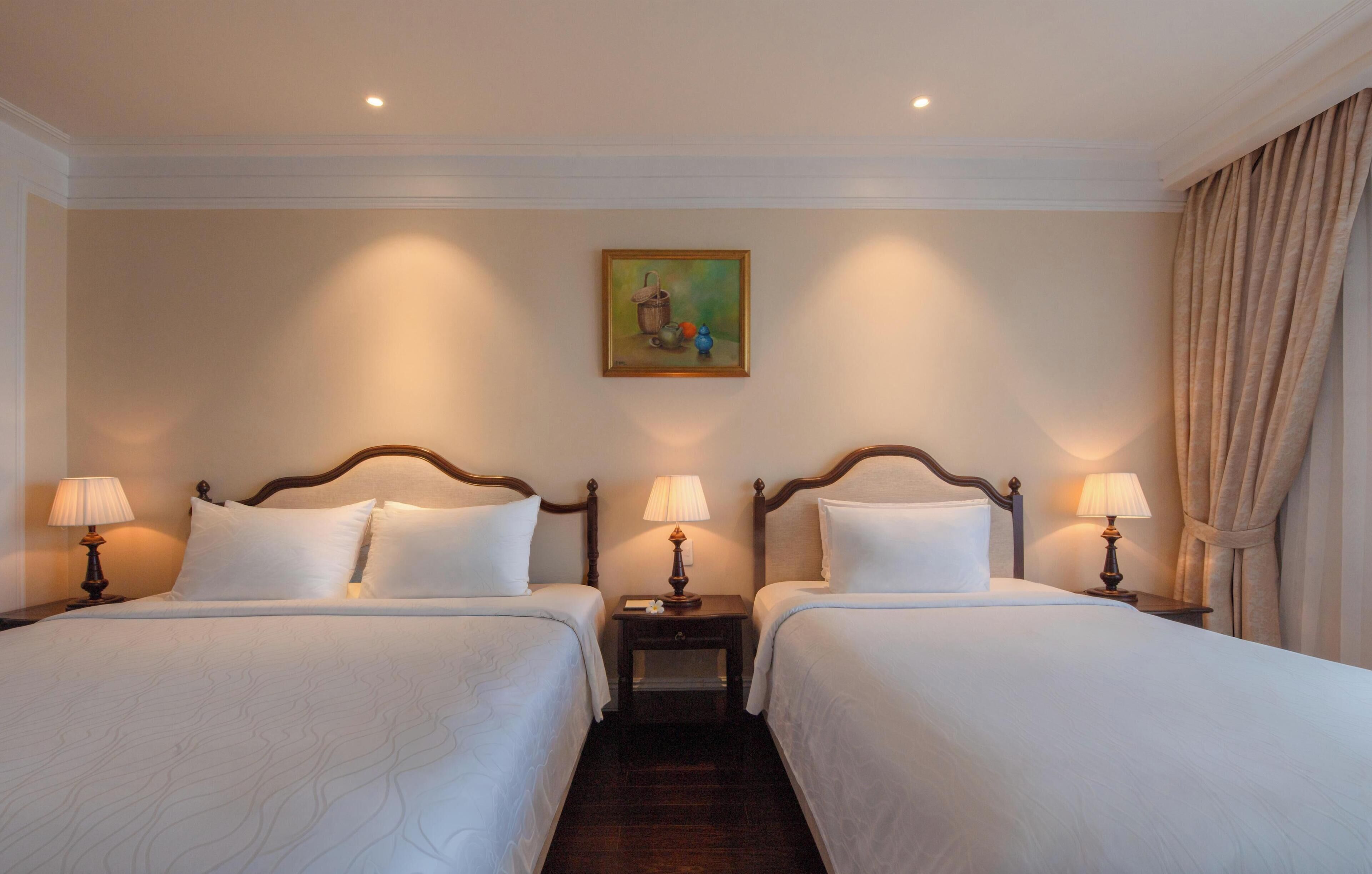 A hotel room with two beds, nightstands with lamps, a painting on the wall, and a window with curtains.