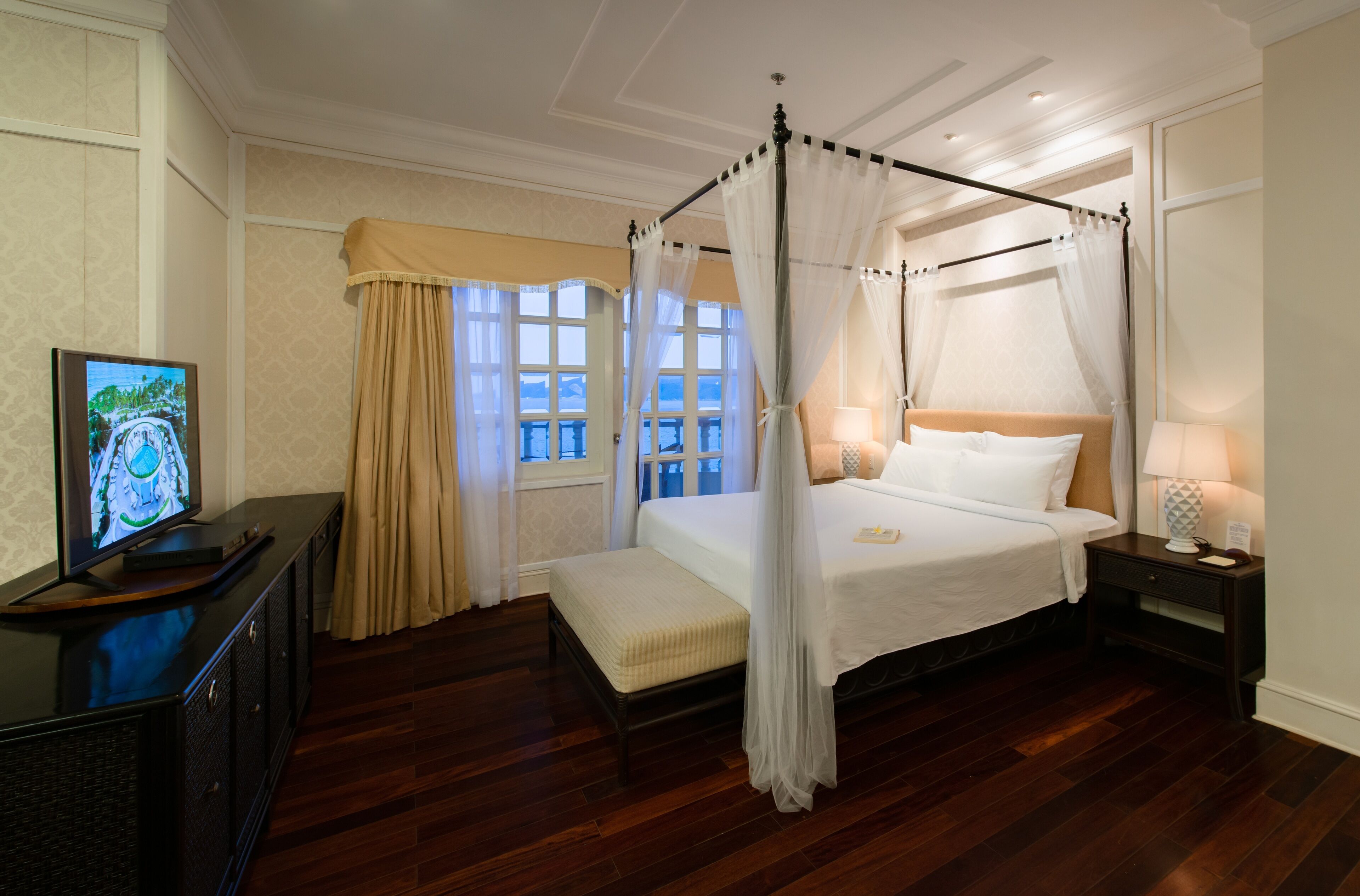 A four-poster bed with a canopy, a bedside table with a lamp, a television on a cabinet, and a window with curtains.