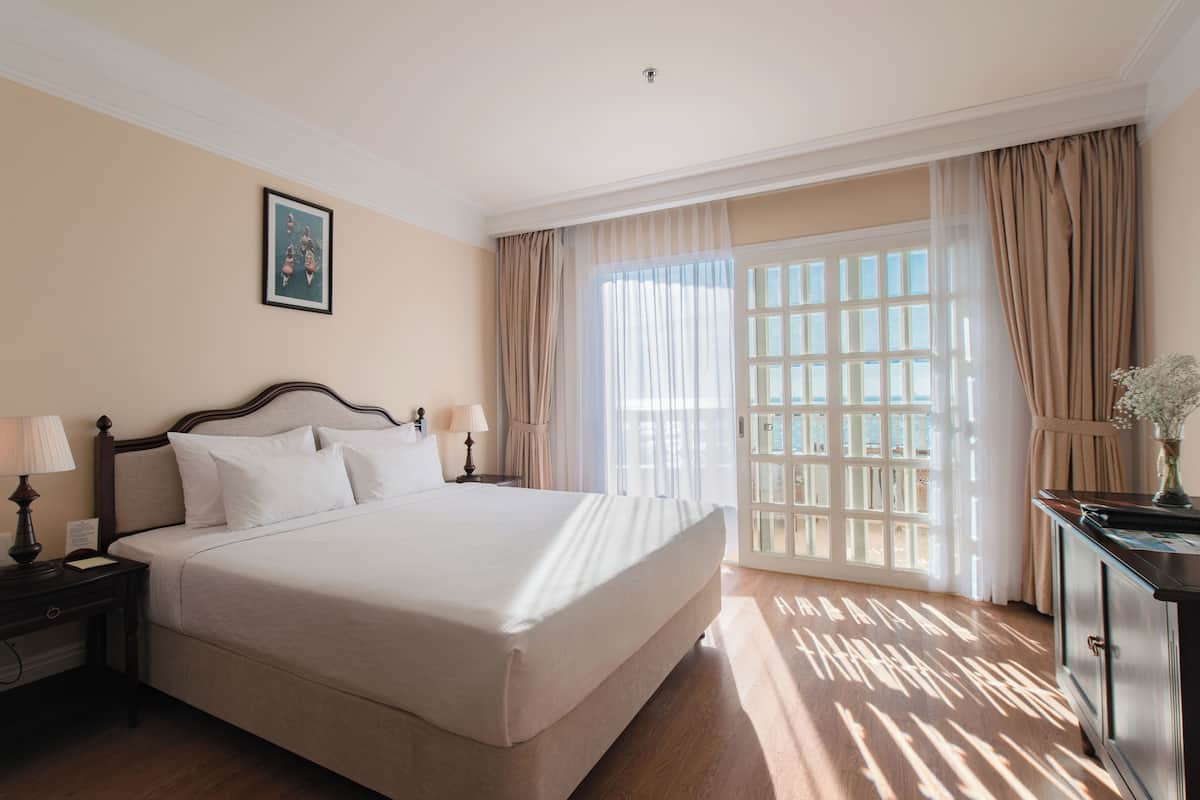 A hotel room with a large bed, two bedside tables with lamps, a nightstand, a dresser, and a large window with curtains.