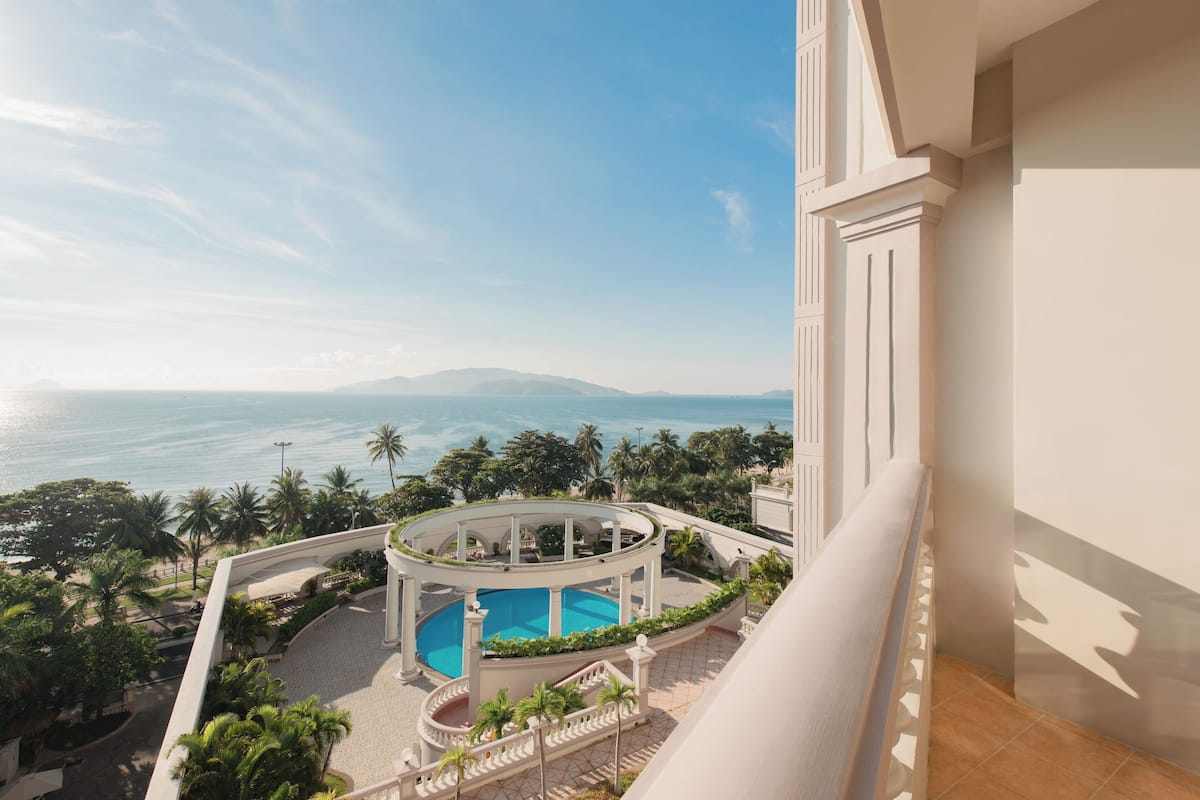 A pool with a view of the sea, surrounded by palm trees and a white building.