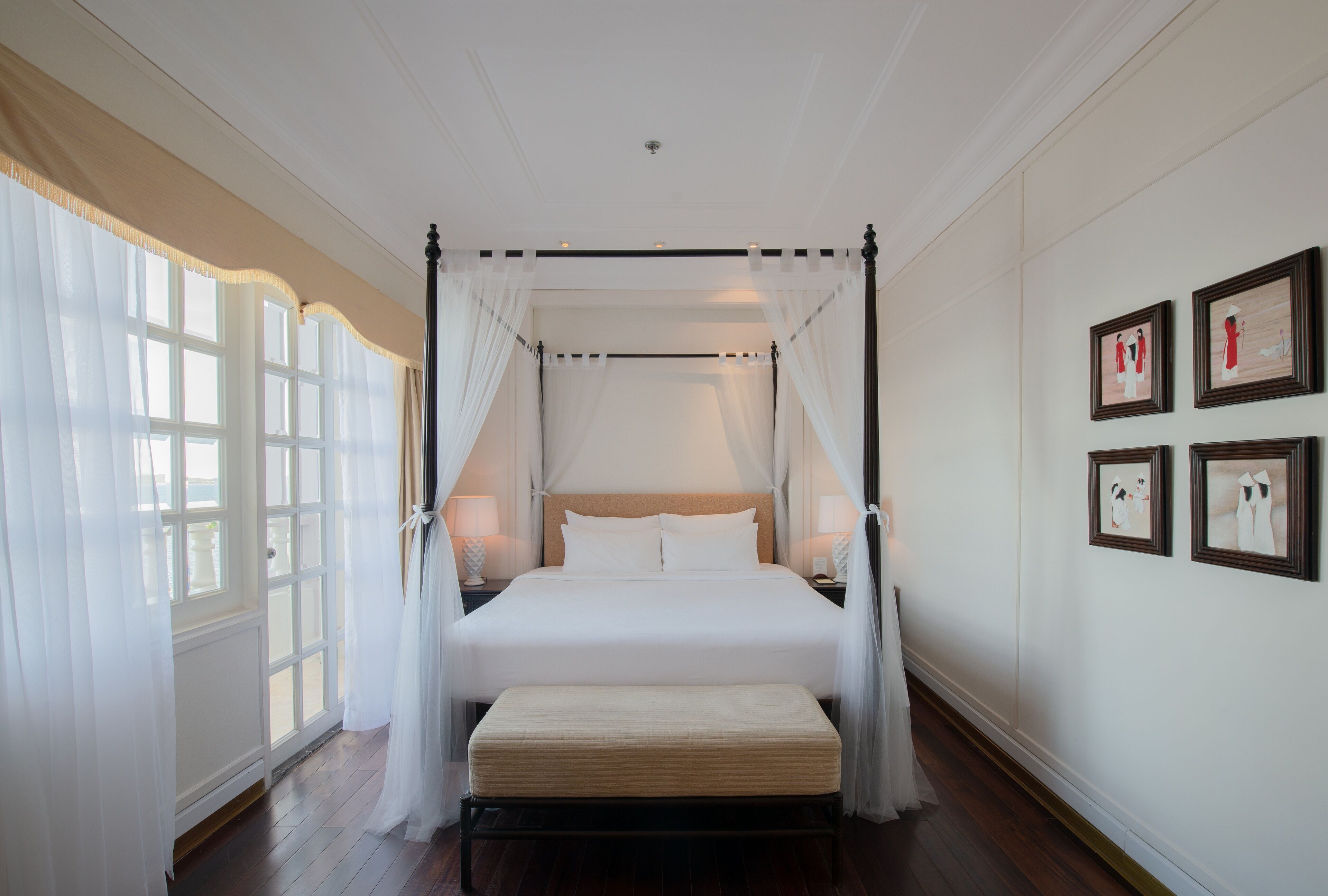 A four-poster canopy bed with white linens, a wooden bench, and a view of the outdoors through large windows.