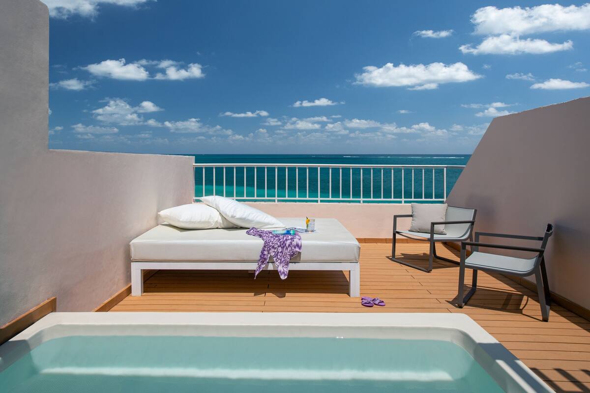 A balcony with a white sofa, a small table, and a chair, overlooking a turquoise sea and clear sky.