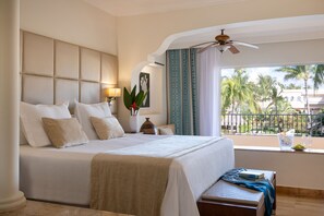 A hotel room with a large bed, a ceiling fan, a view of palm trees, and a balcony with a railing.