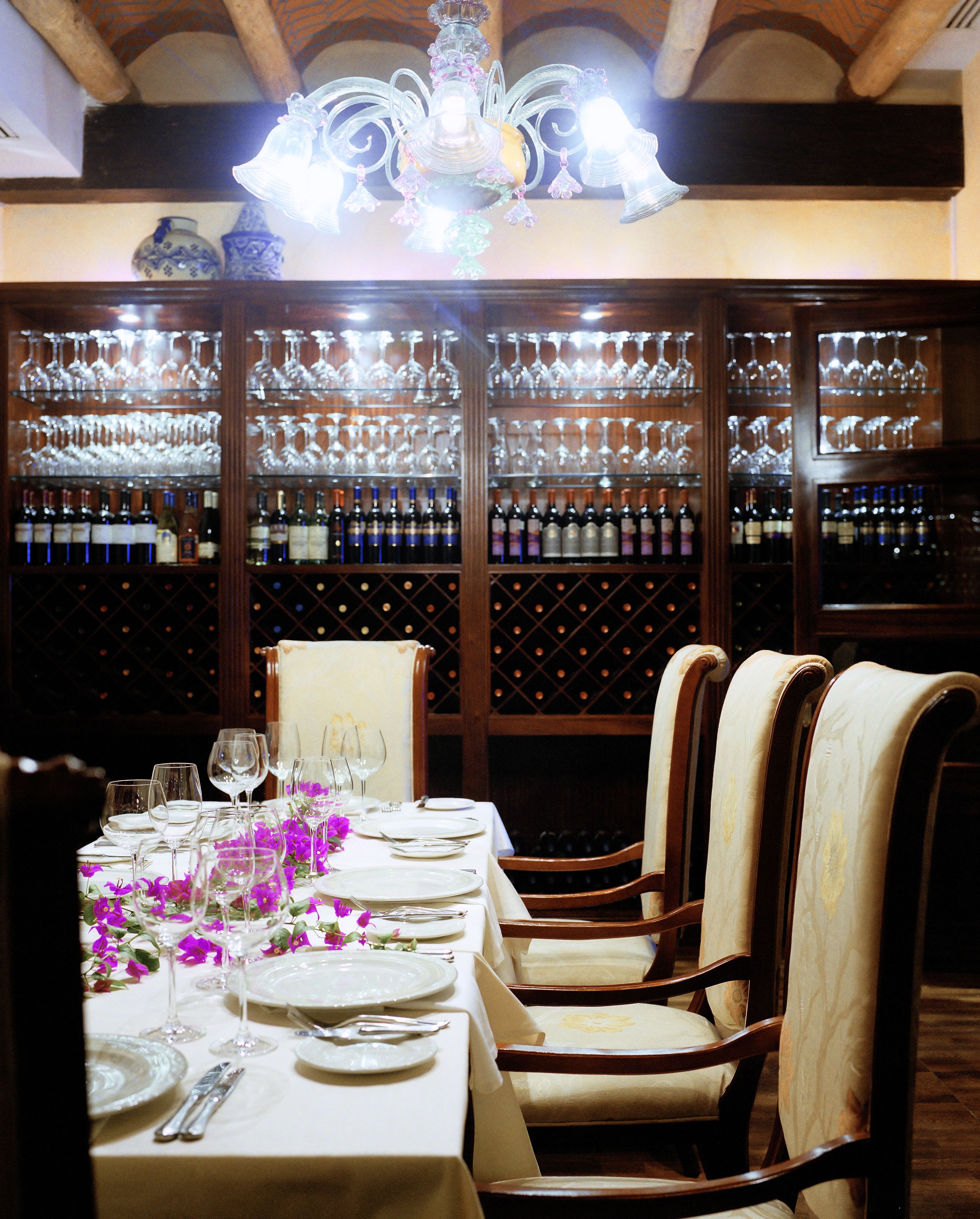 Un comedor con una mesa larga dispuesta para una comida, rodeada de sillas de madera. Detrás de la mesa, hay una estantería para vinos con varias botellas.