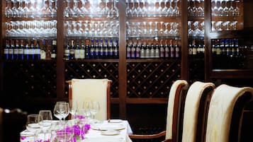 Un comedor con una mesa larga dispuesta para una comida, rodeada de sillas de madera. Detrás de la mesa, hay una estantería para vinos con varias botellas.
