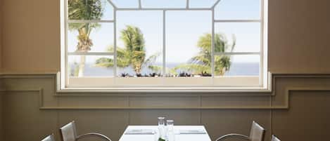 A dining area with a table set for six, a large window with a view of palm trees and the ocean, and a neutral color scheme.