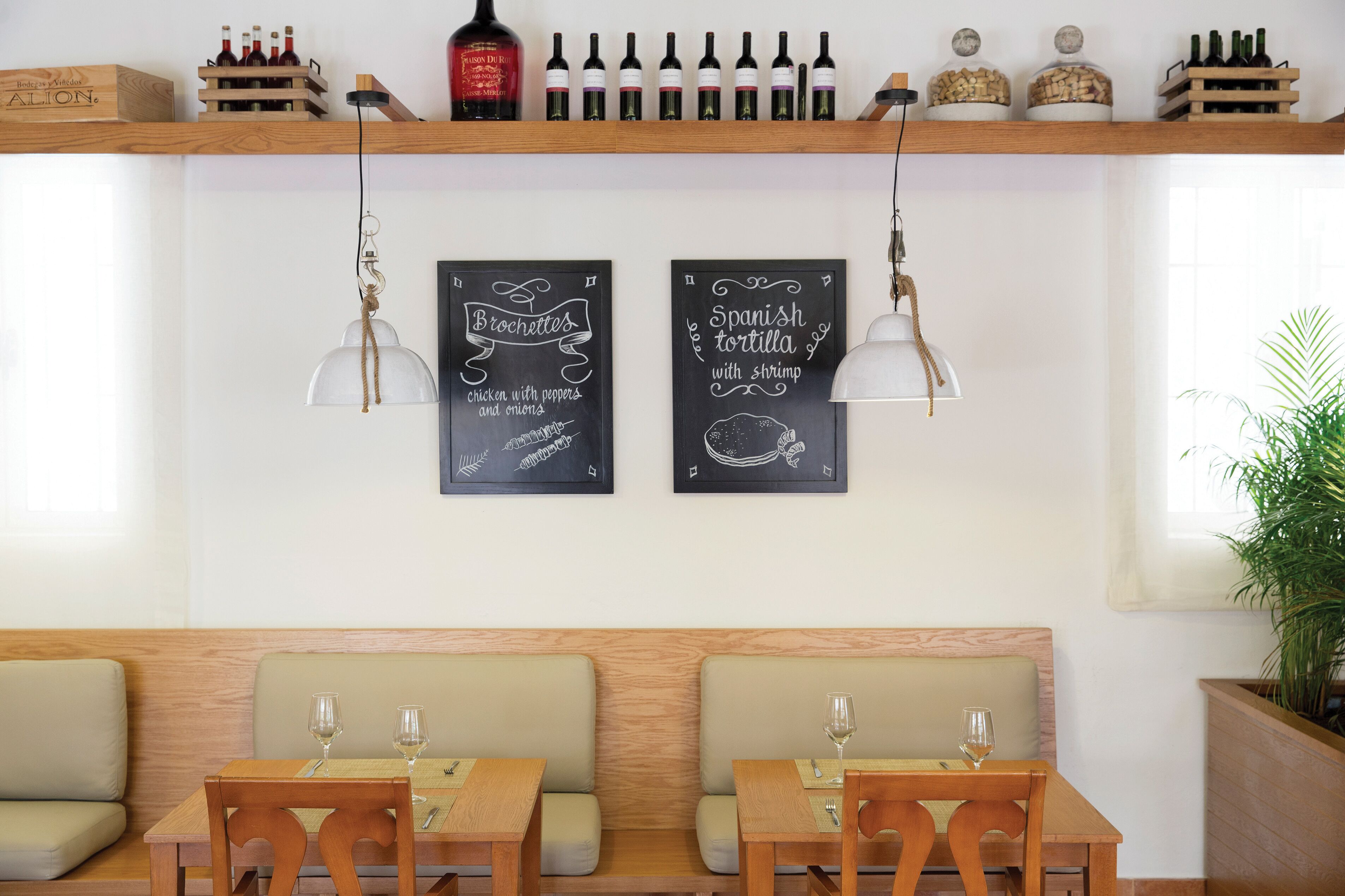 Un restaurante con dos mesas y bancos, botellas de vino en estanterías y dos carteles de pizarra con productos alimenticios.
