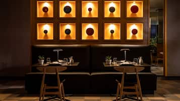 Un restaurante con interior oscuro, un separador de madera con decoraciones circulares iluminadas, un sofá de cuero negro y dos mesas de madera con sillas.