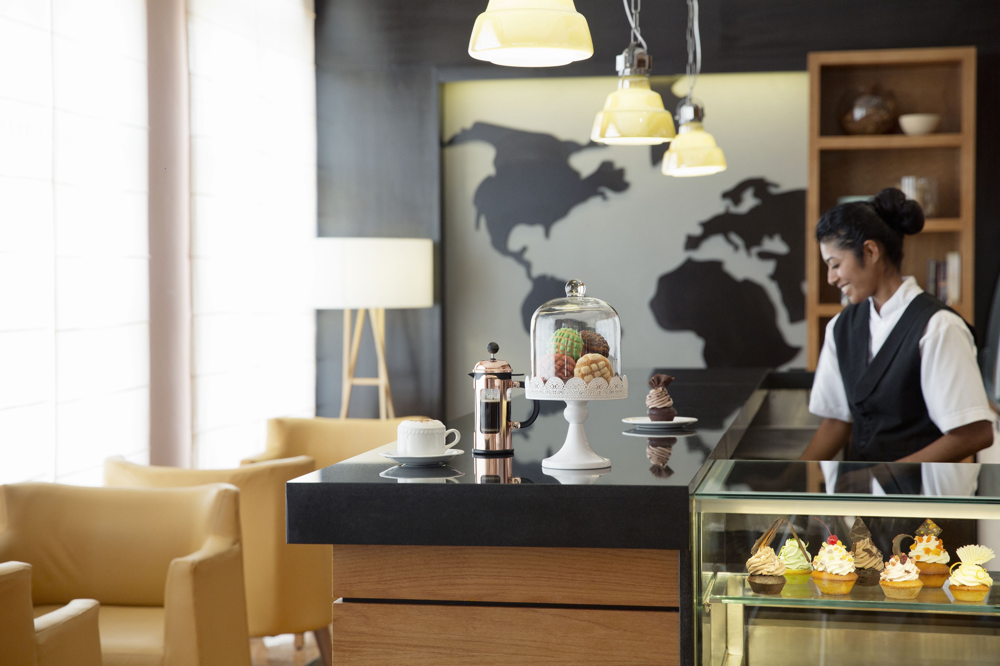 Interior de una cafetería moderna con una máquina de café, una vitrina con pasteles y un barista trabajando.