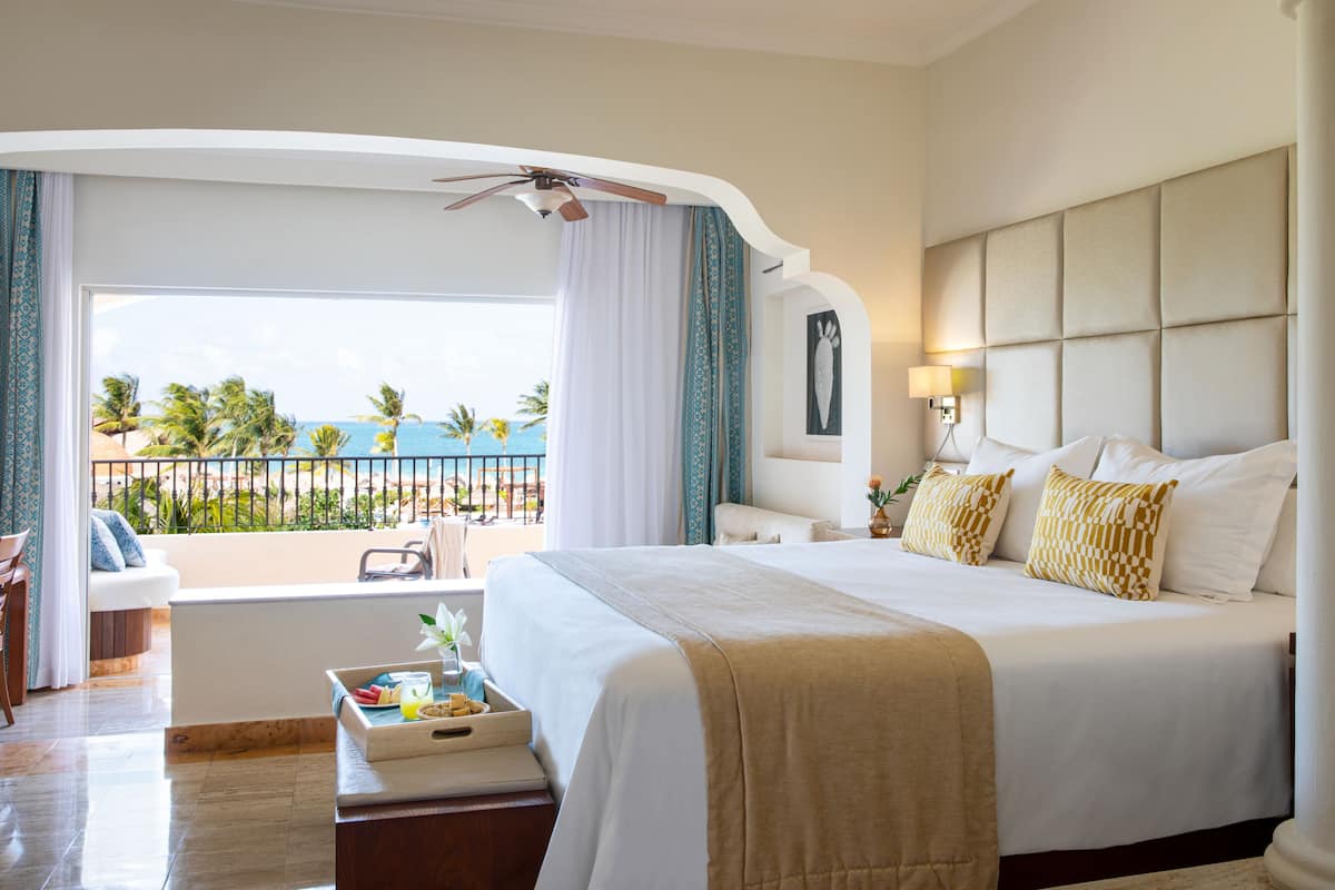A hotel room with a large bed, a balcony with a view of the beach, and a small seating area with a table.