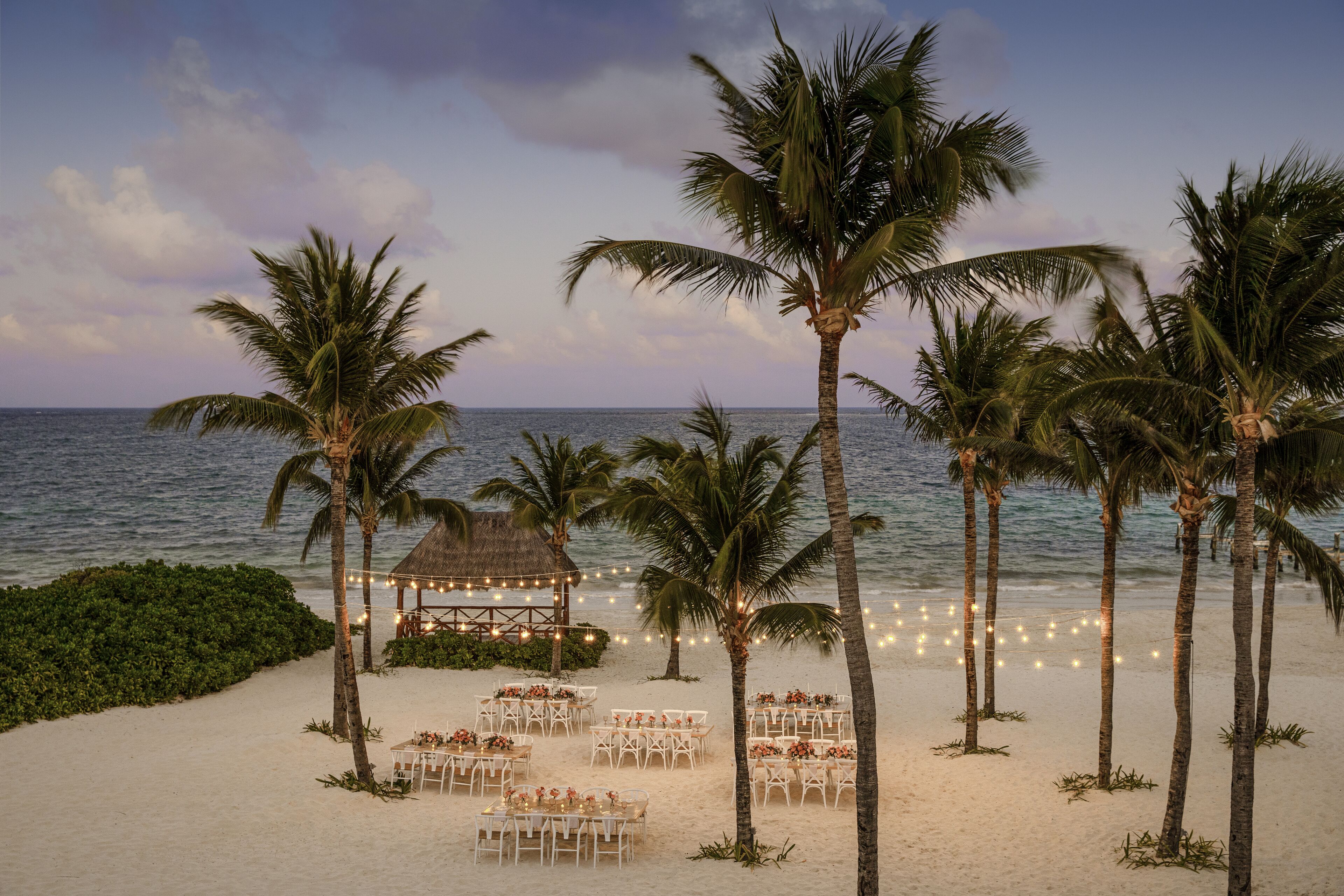 Preparação para um casamento na praia, com mesas, cadeiras, palmeiras e luzes de fada.