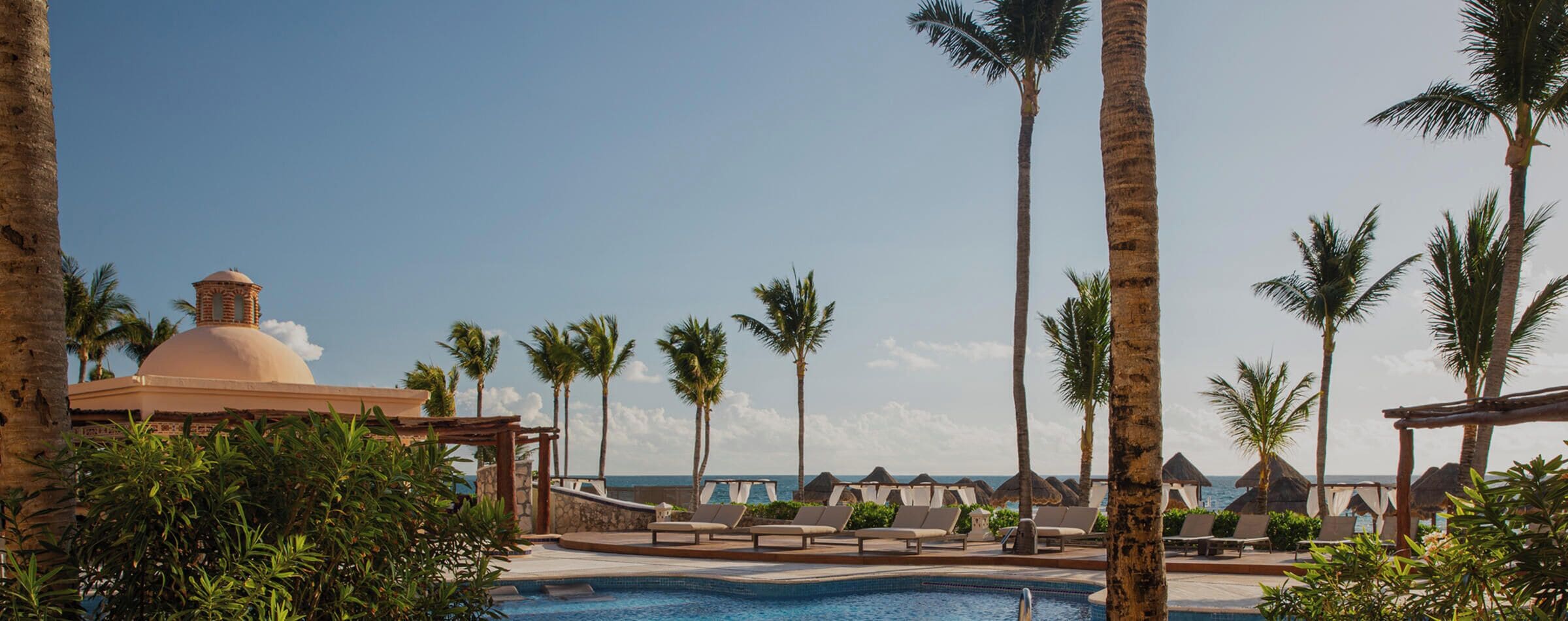 Un área de piscina con sillones de descanso, palmeras y un edificio con cúpula.