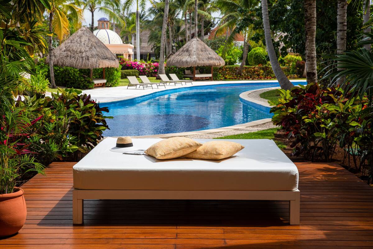 A poolside lounge area with a wooden deck, a white lounge chair, and a thatched umbrella.