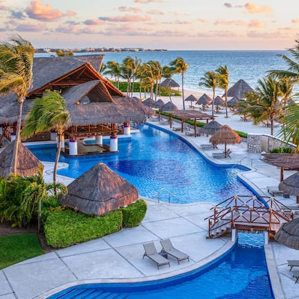 A resort pool area with a view of the ocean, surrounded by thatched umbrellas and palm trees.