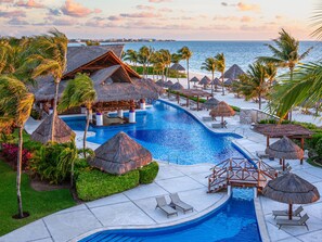 A resort pool area with a view of the ocean, surrounded by thatched umbrellas and palm trees. - Excellence Riviera Cancun - Adults Only All Inclusive (Puerto Morelos)