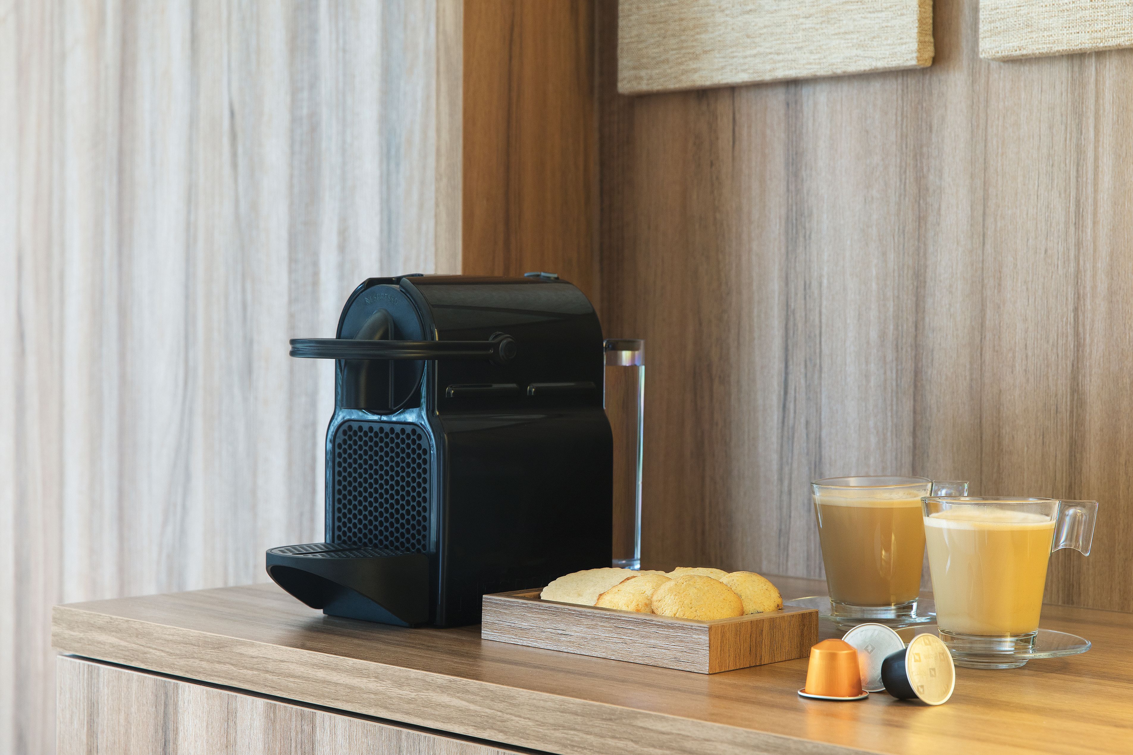 A coffee maker, two glasses of coffee, a tray of cookies, and a cup of tea on a wooden table.