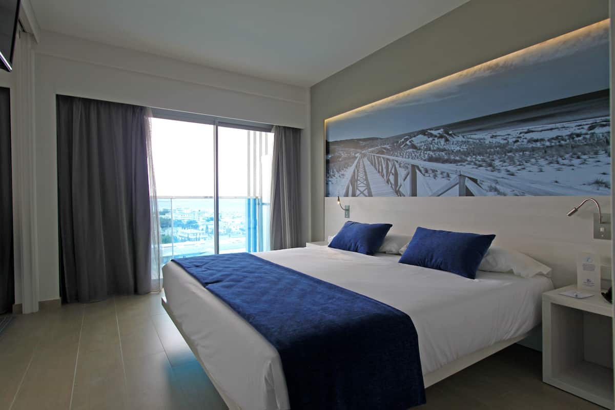 A modern hotel room with a large bed, a nightstand, a painting of a bridge, and a view of a snowy landscape through the window.