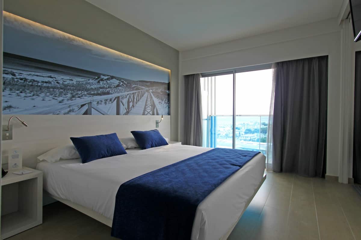 A modern hotel room with a large bed, a nightstand, a painting of a bridge, and a view of a cityscape through the window.