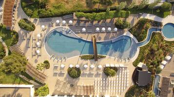 Vista aérea de un complejo turístico con dos piscinas grandes, tumbonas y zonas con sombra.