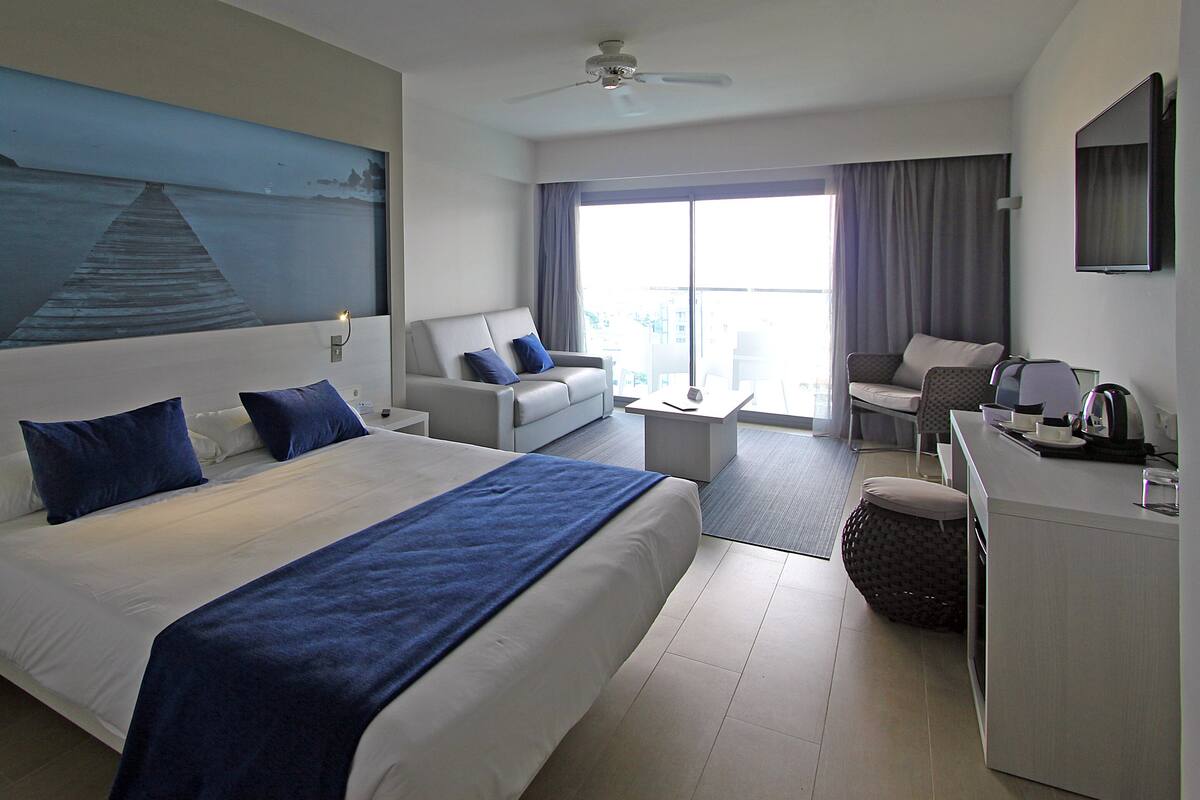 A hotel room with a large bed, a desk, a chair, a sofa, a TV, and a painting of a seascape.