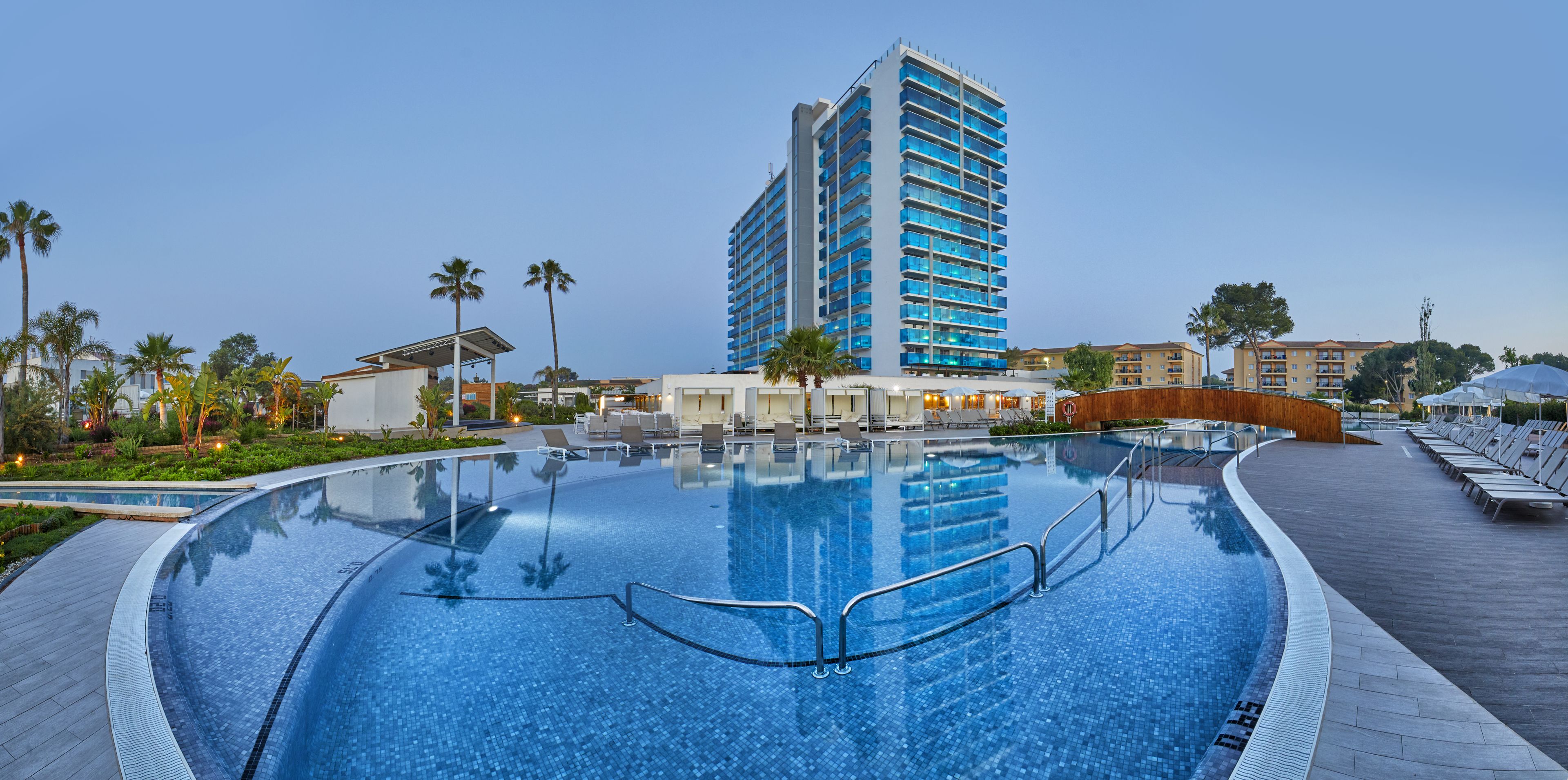 A modern hotel with a large pool, palm trees, and a tall building in the background.