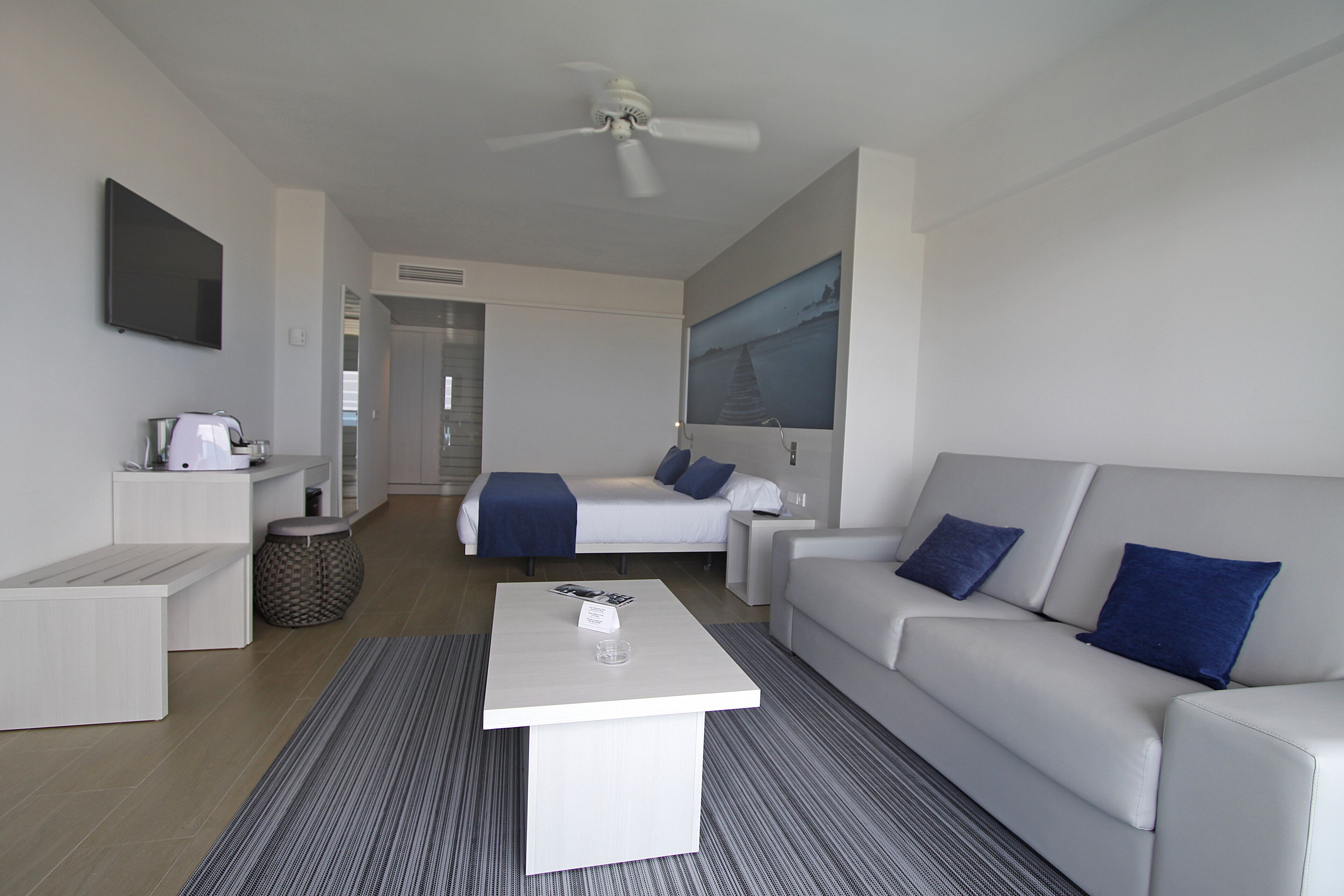 A modern hotel room with a white sofa, a coffee table, a bed with blue pillows, a flat-screen TV, and a large window.