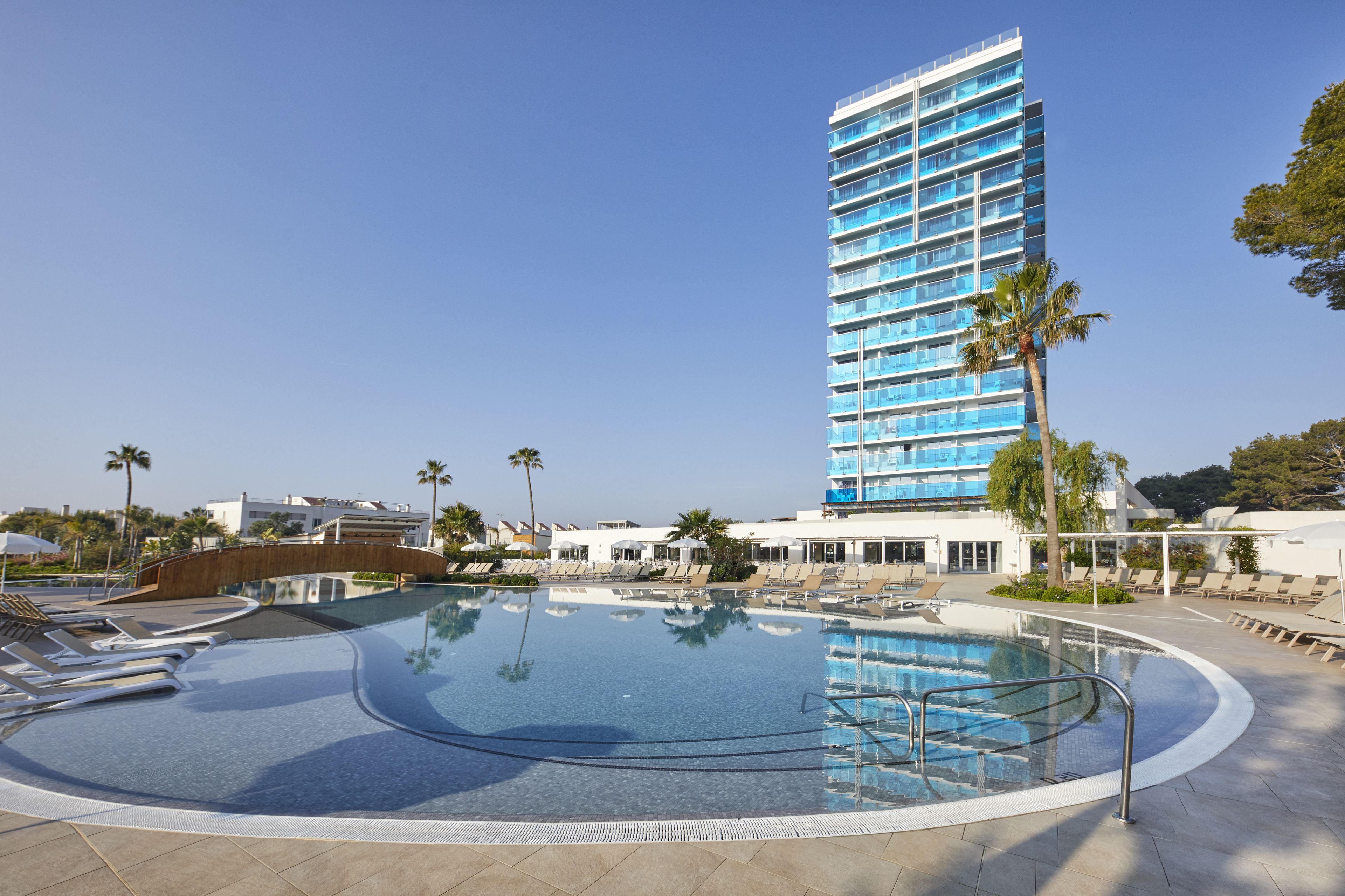 A modern hotel with a large pool area, palm trees, and a clear blue sky.